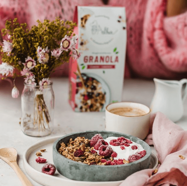 een ontbijt met granola, yoghurt, frambozen en koffie en op de achtergrond een vaasje met bloemen